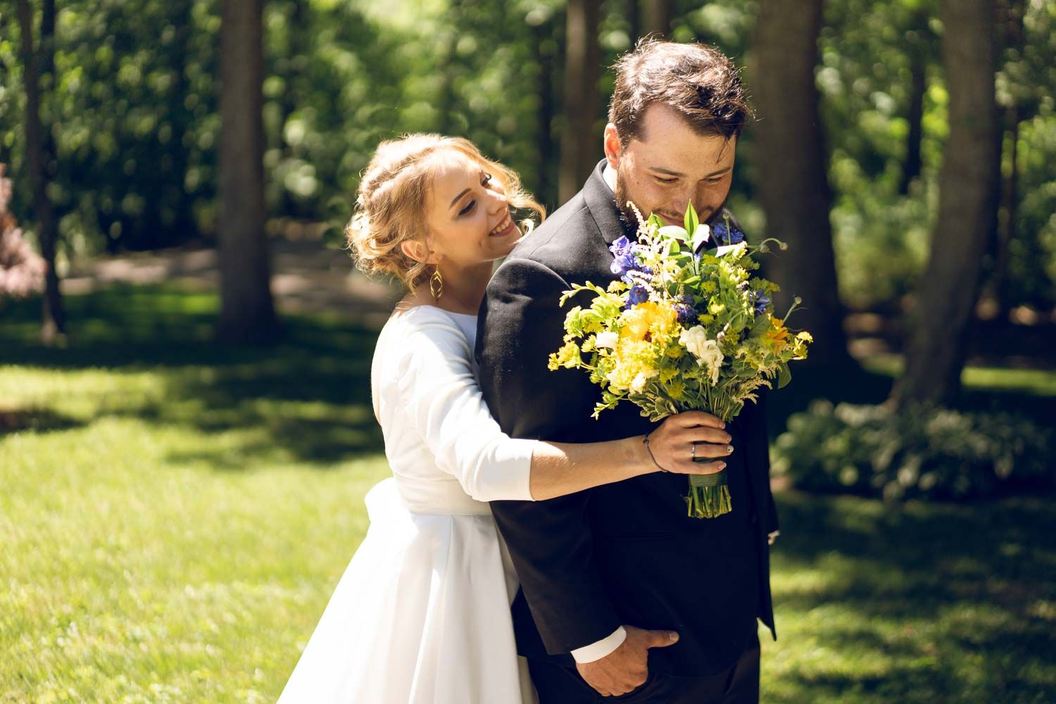 Bride hugging groom from behind while holding colorful bouquet in lush green park setting.