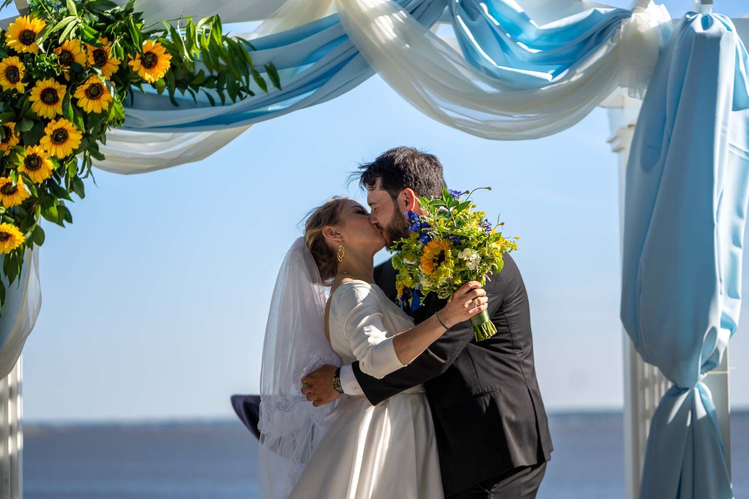 Bride and groom share first kiss under sunflower-adorned arch with blue sky and water backdrop.