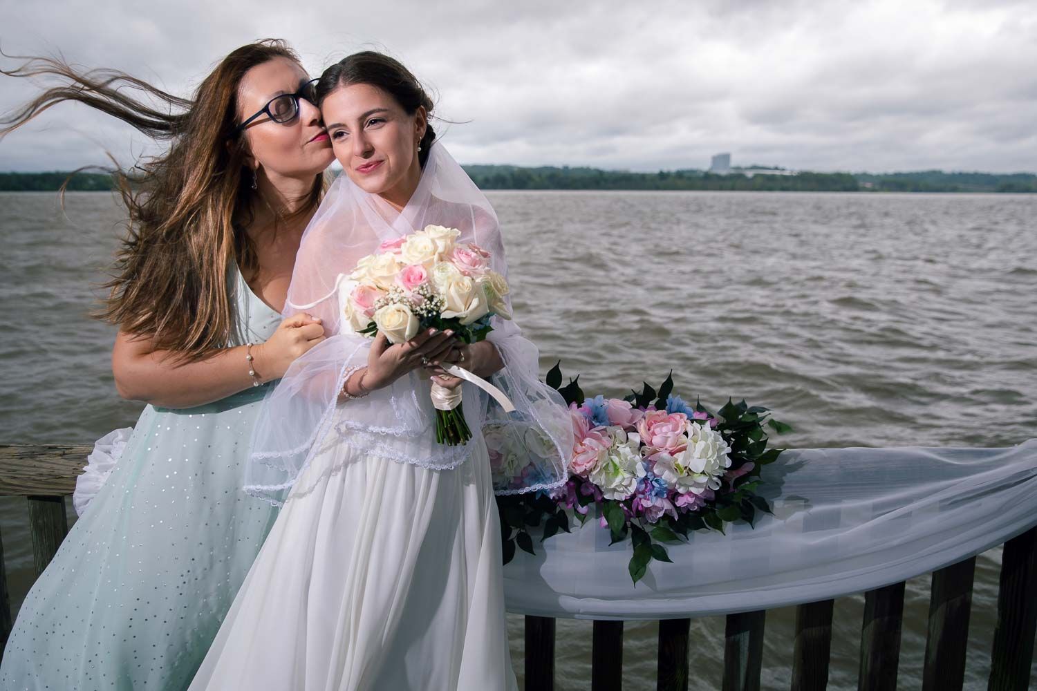Two women posing by waterfront railing, bride in white gown holding bouquet on windy day.