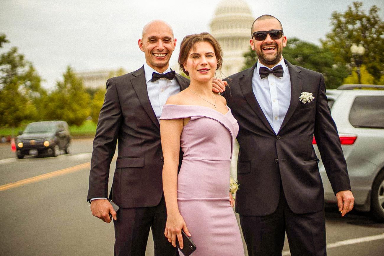 Two men in tuxedos and a woman in lavender dress posing outdoors near US Capitol building.