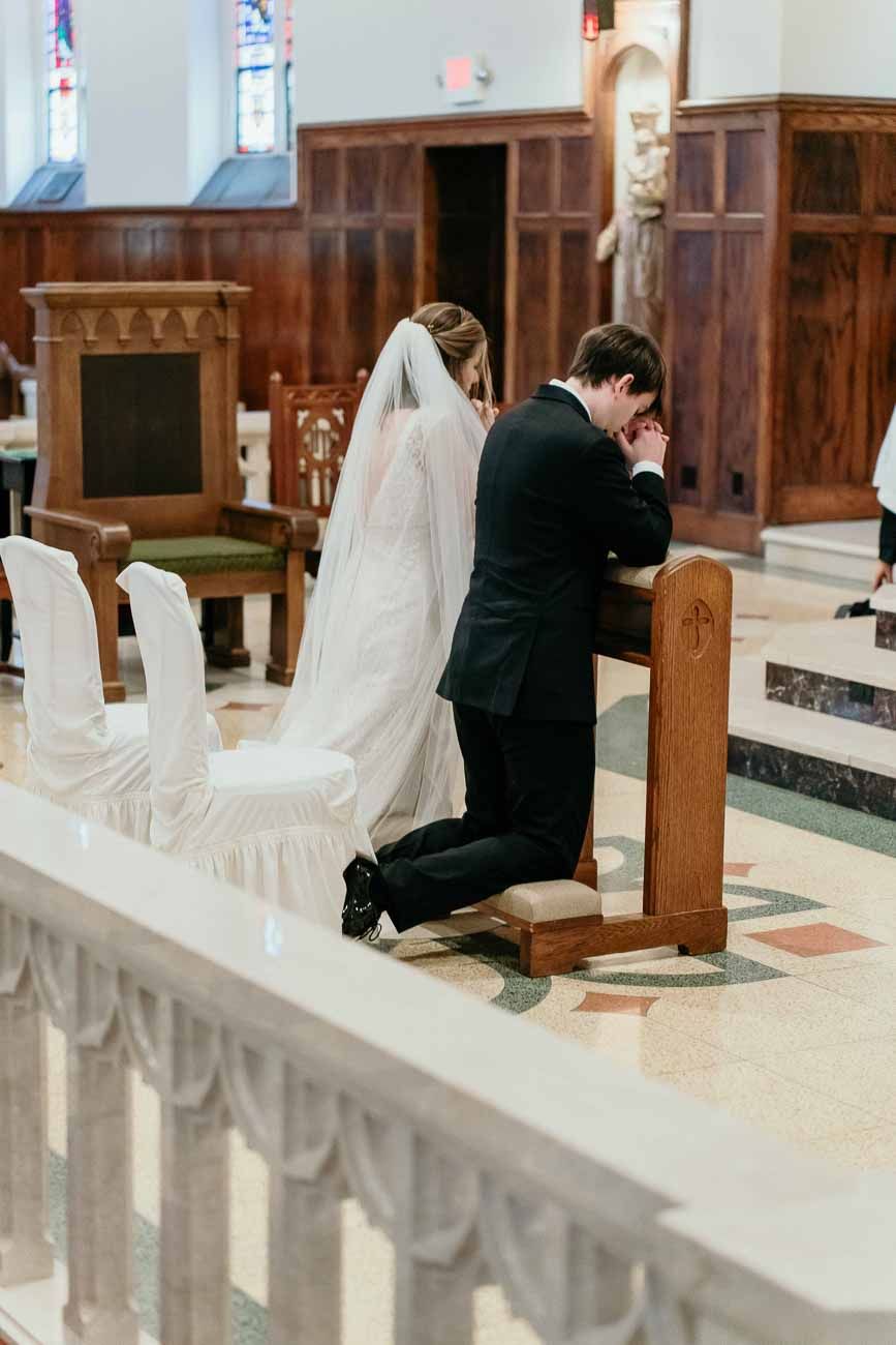 Bride in white veil and groom in black tux kneel together in prayer at a Catholic church wedding.