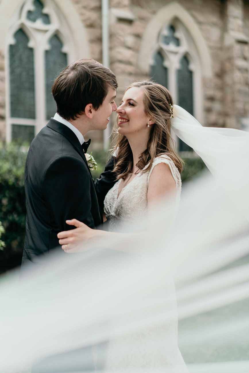 Smiling bride gazes at groom while her veil billows in foreground outside a Gothic stone church.