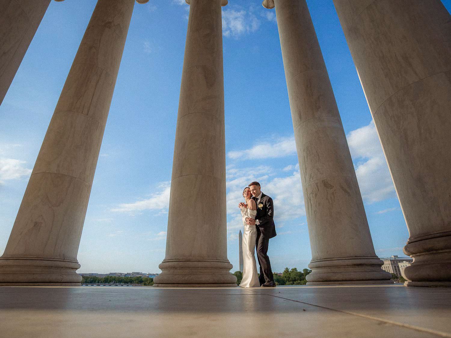 Bride and groom embrace between tall marble columns at the Jefferson Memorial under a blue sky.