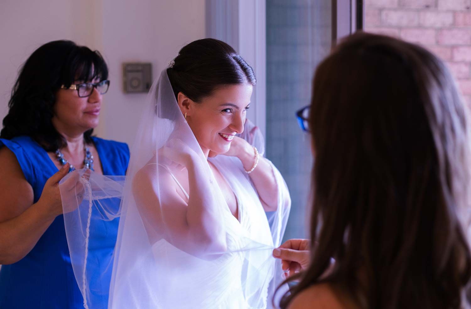 Bride adjusting her veil while smiling, assisted by bridesmaids in a bright room during wedding preparations.