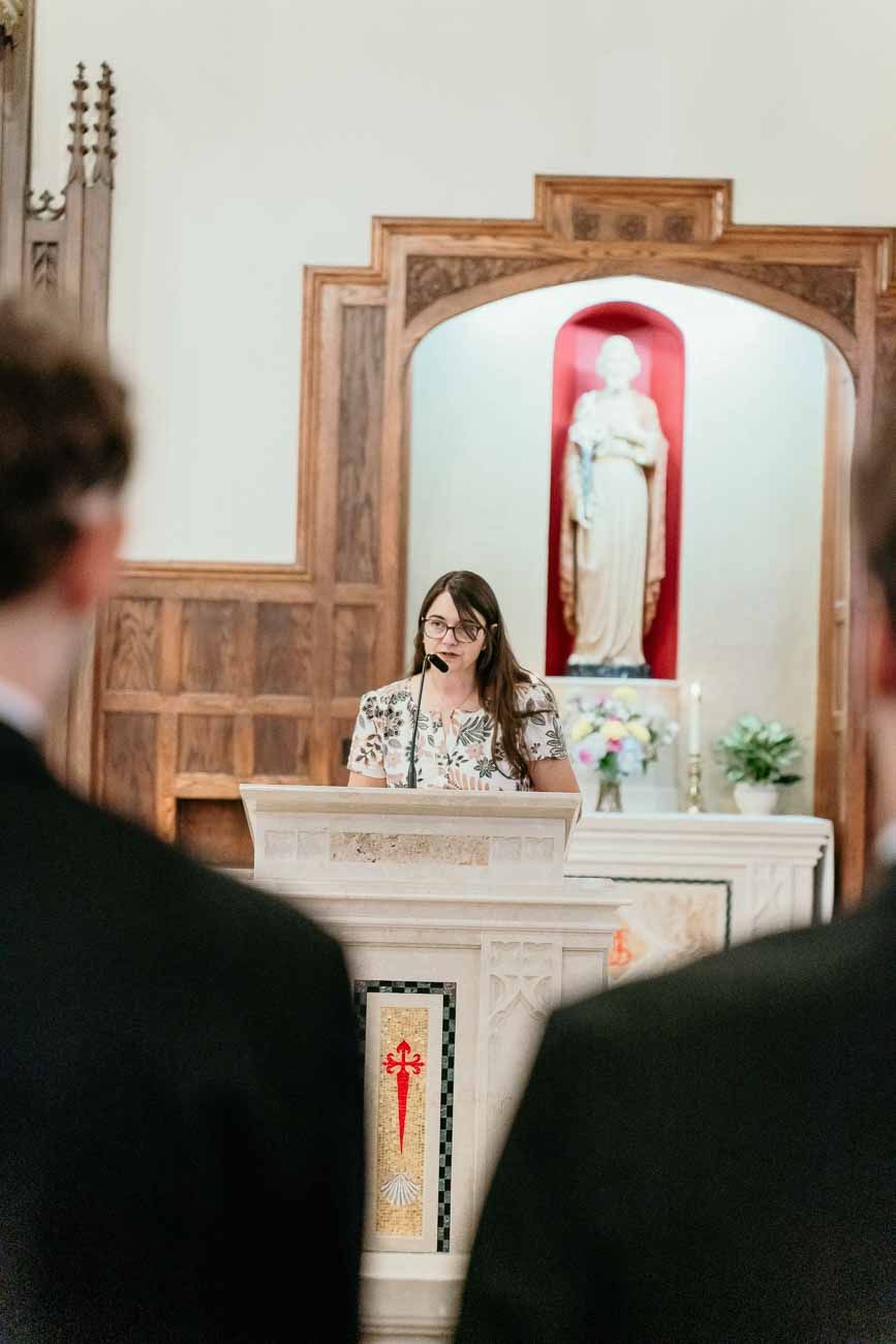 Young woman in floral dress delivers a reading at a church podium during wedding ceremony.