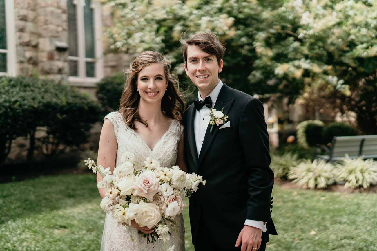 Smiling bride and groom pose outdoors holding a white and blush floral bouquet near a stone church.
