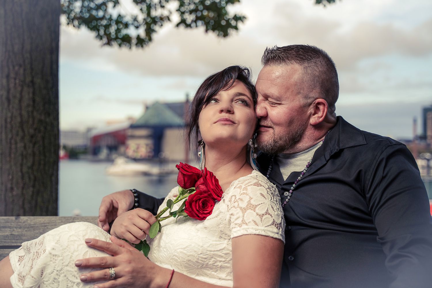 Couple sharing a romantic moment by the waterfront, woman in lace dress holding red roses, man in black shirt embracing her.