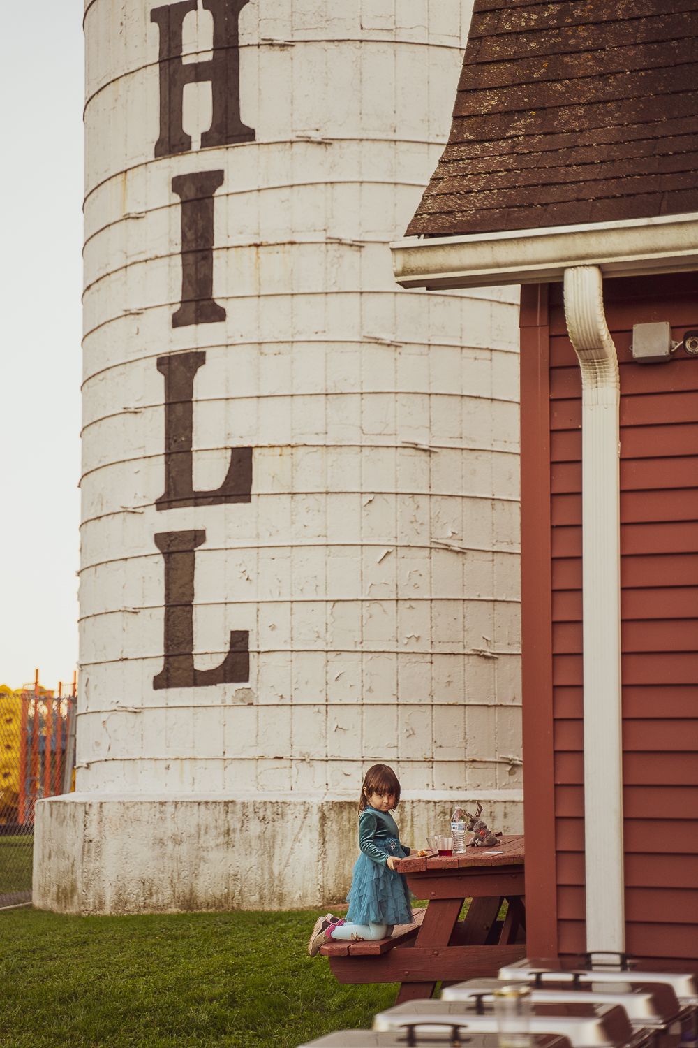 Young girl in blue dress standing beside a large white grain silo at a farm venue.