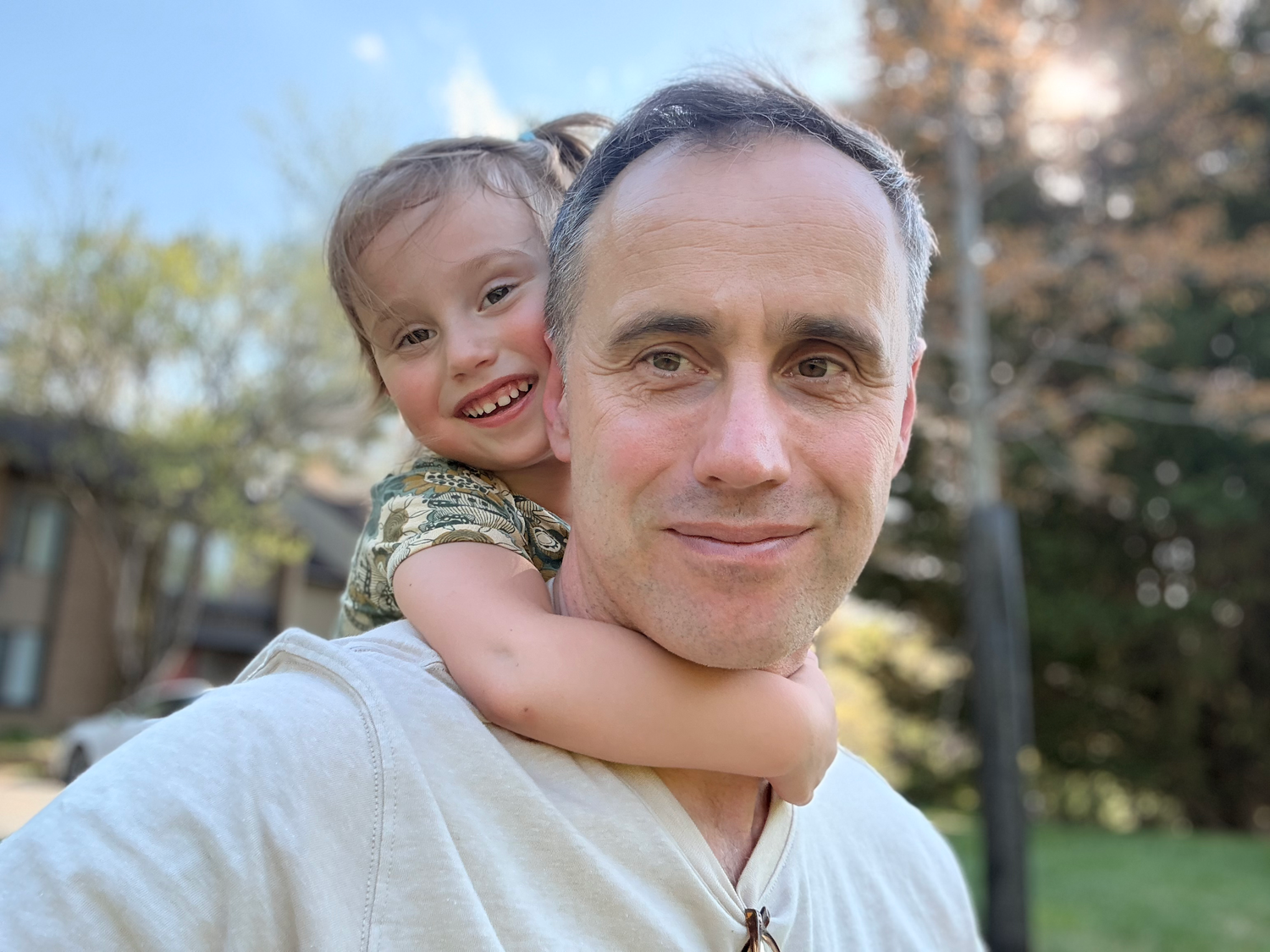 Smiling father giving his laughing young child a piggyback ride outdoors in a sunny backyard setting.