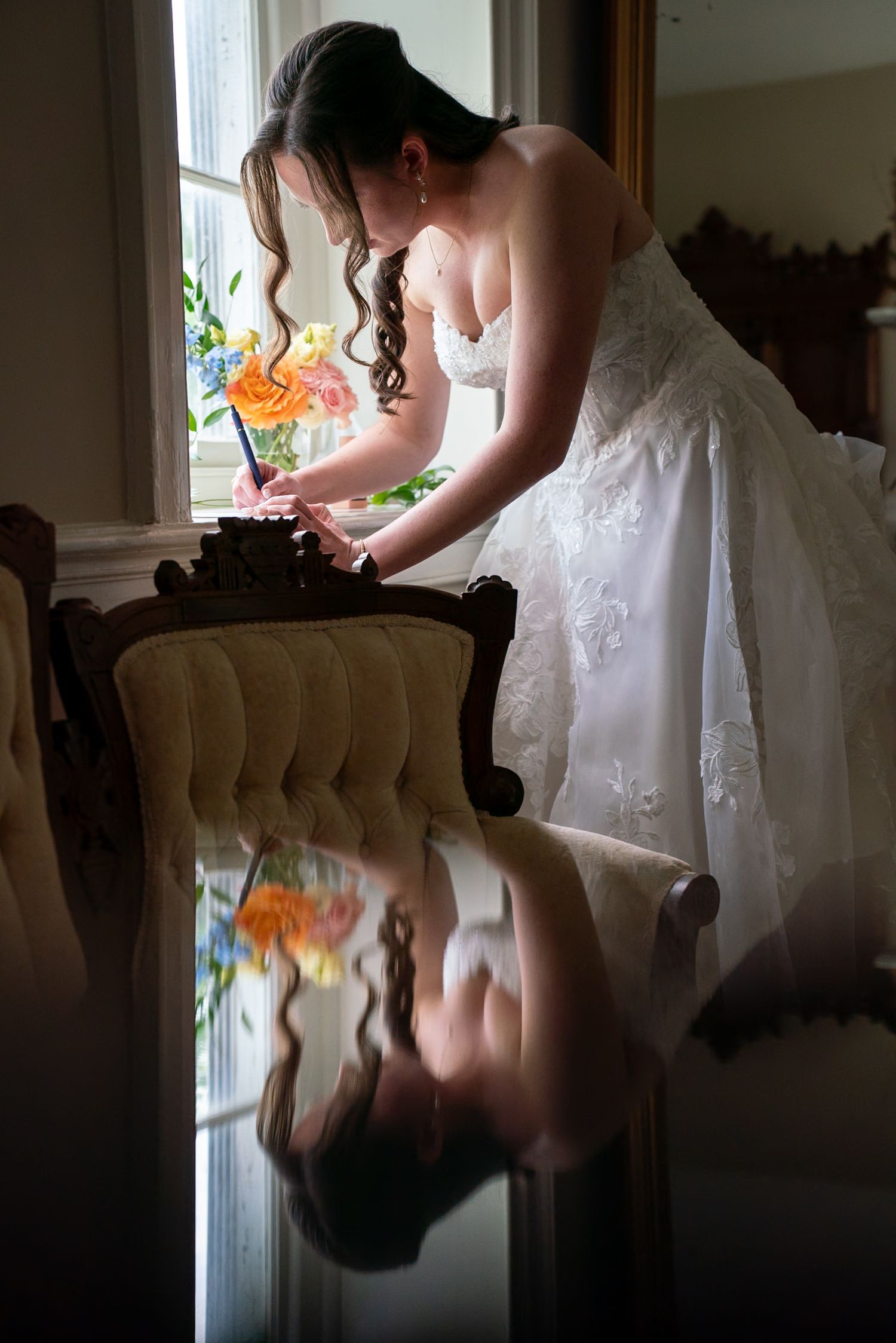 Bride in white gown writing at antique chair by window, reflection visible on glossy floor surface.