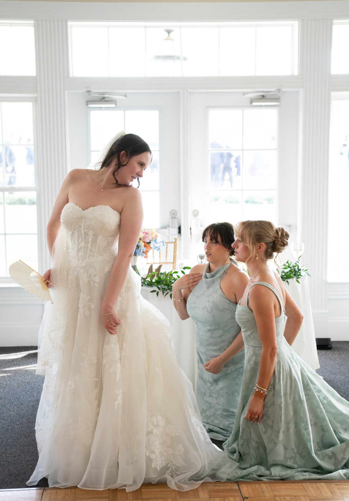 Bride in strapless white lace gown with two bridesmaids in sage green dresses adjusting her dress indoors.