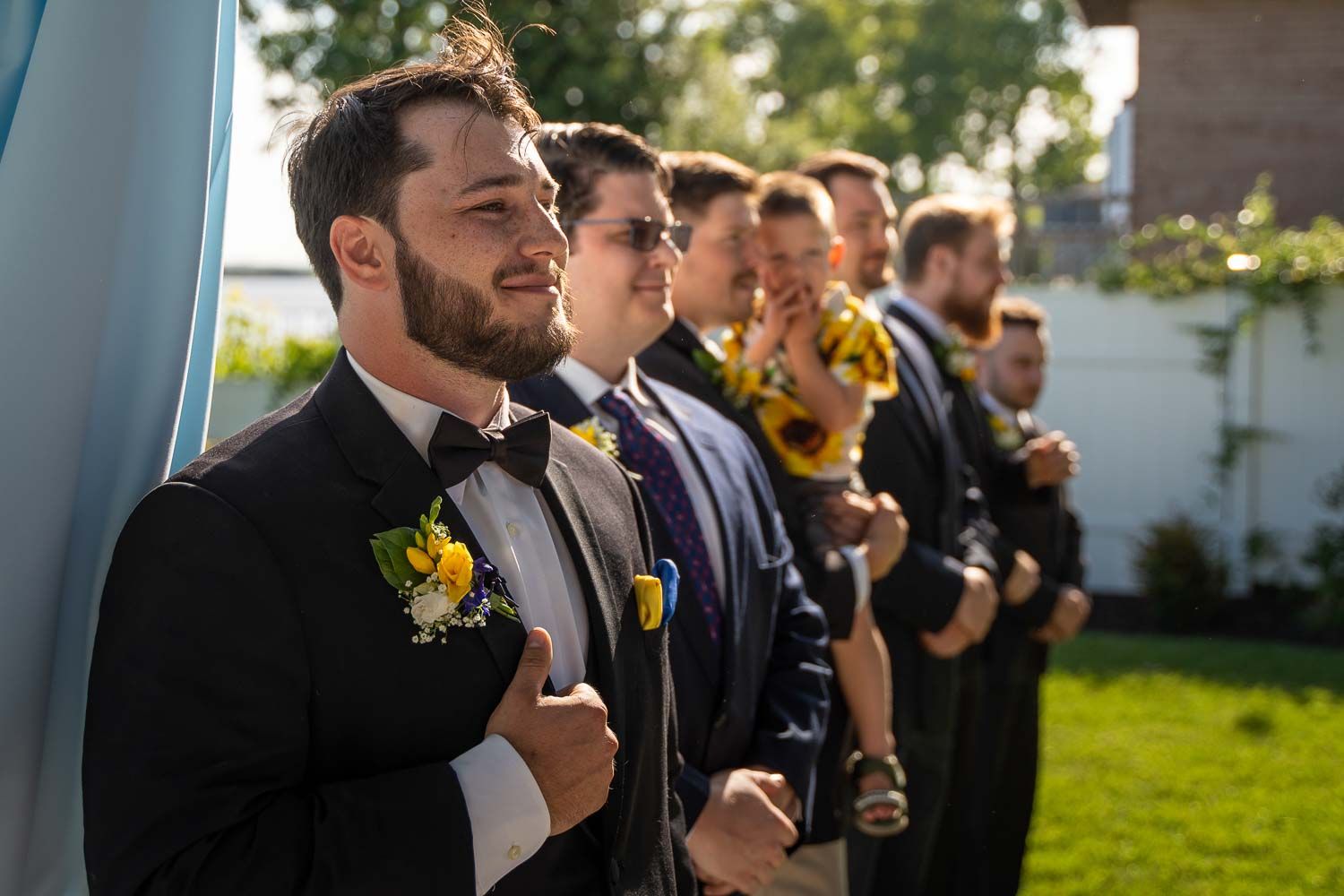 Groom in tuxedo with yellow boutonniere stands with groomsmen in navy suits at outdoor wedding ceremony.
