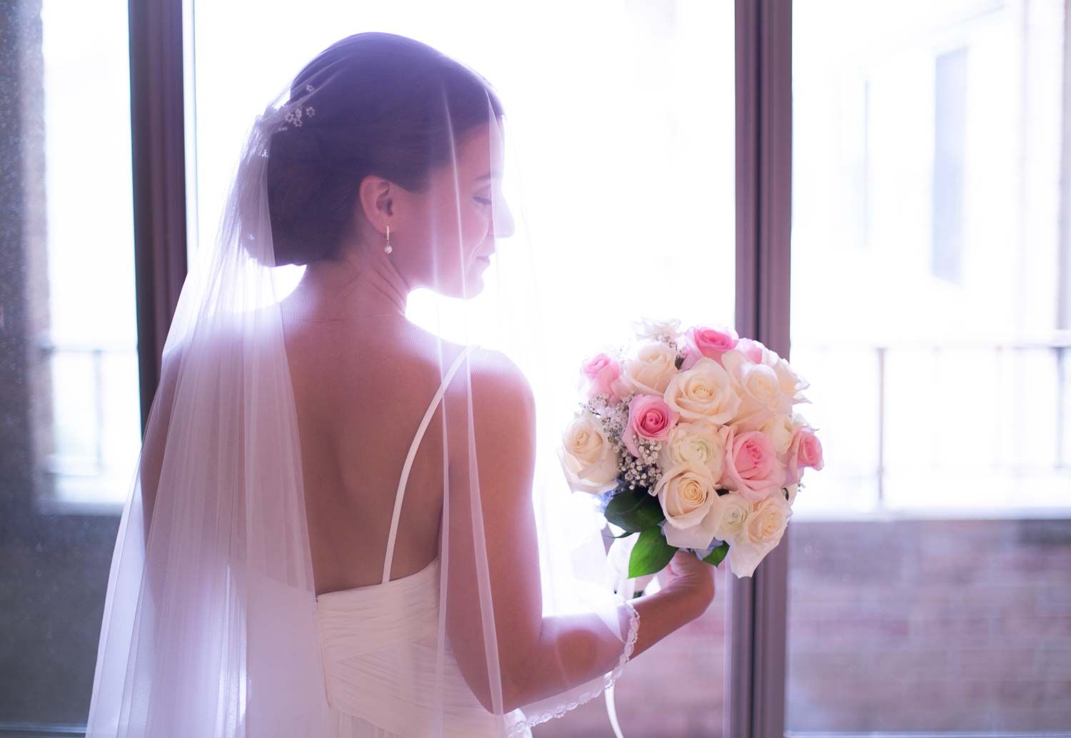 Elegant bride in white gown and veil holding pink and white rose bouquet while gazing out a bright window.