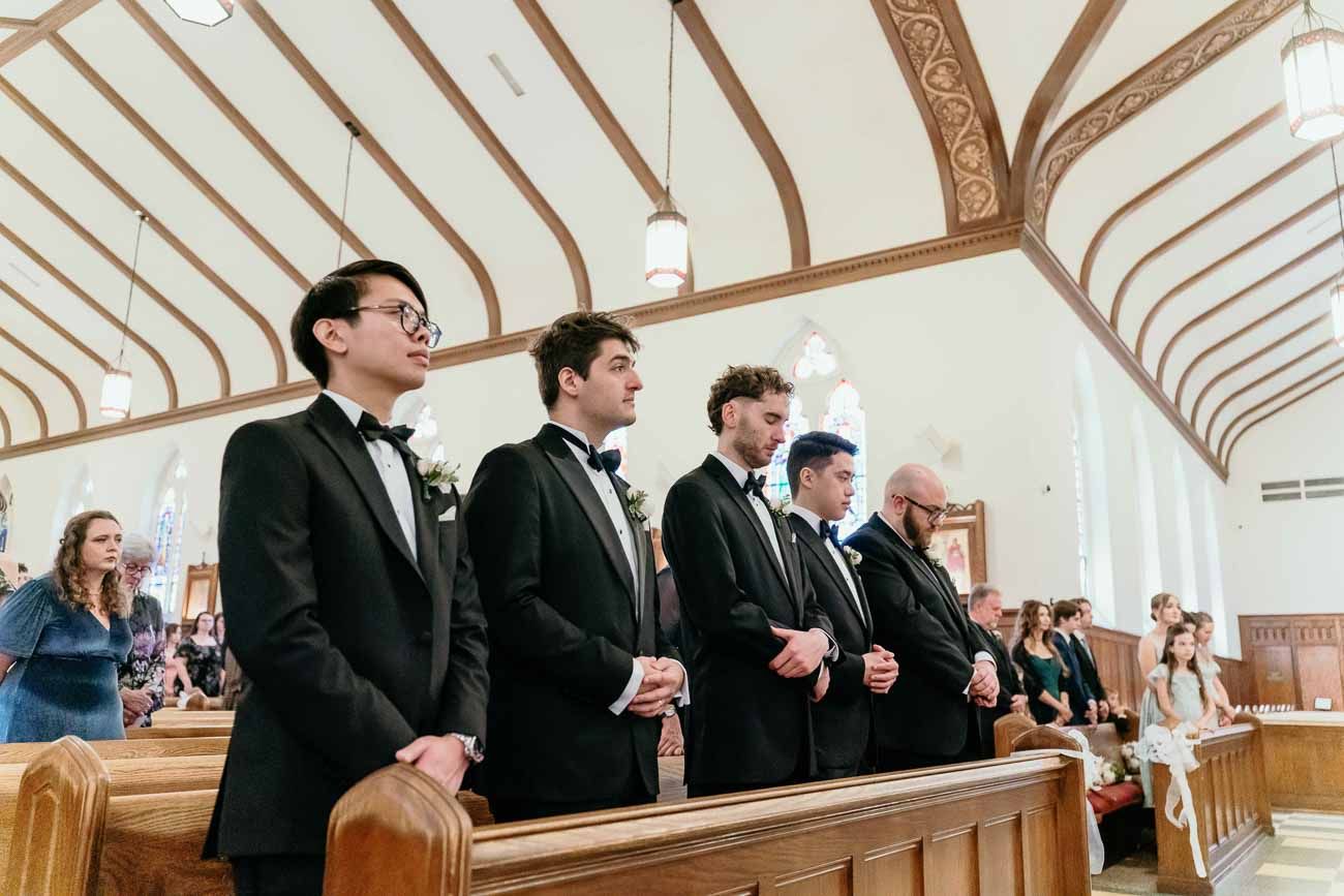 Groomsmen in black tuxedos standing at attention in church pews during wedding ceremony.