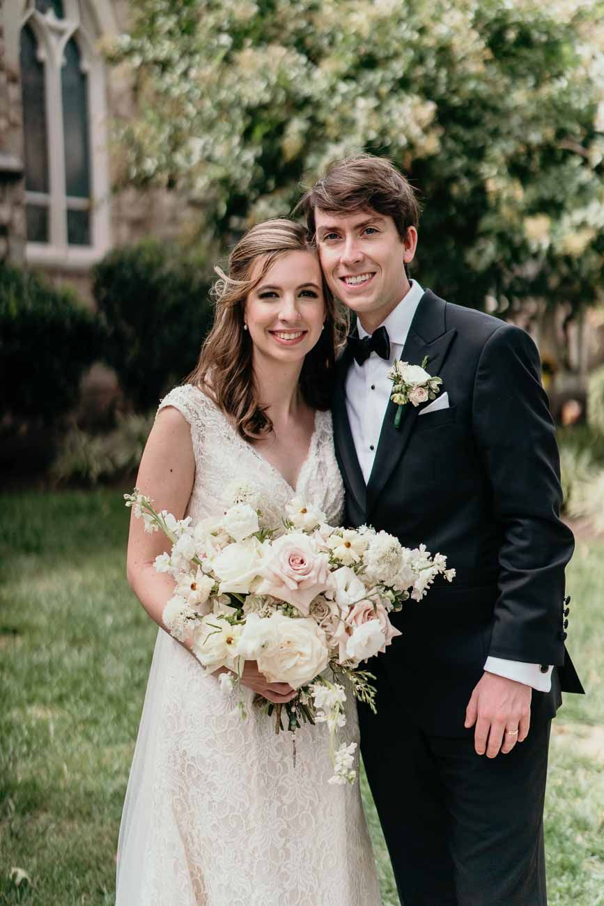 Happy bride and groom pose together outdoors with a lush white and pink bridal bouquet near a church.