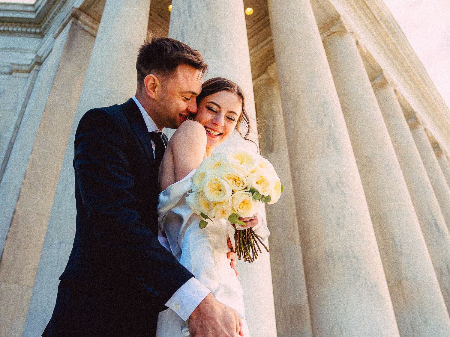 Groom embraces bride holding white rose bouquet beside towering marble columns at the Jefferson Memorial.