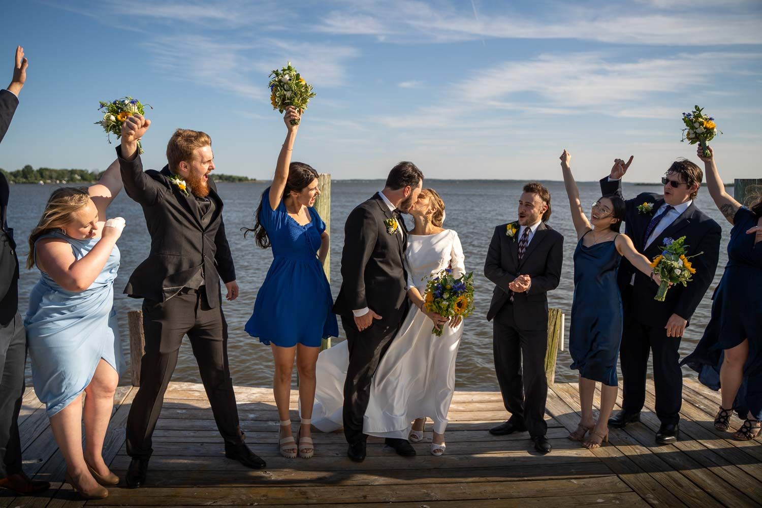 Wedding party celebrating on lakeside dock, bridesmaids in blue dresses raising bouquets joyfully.