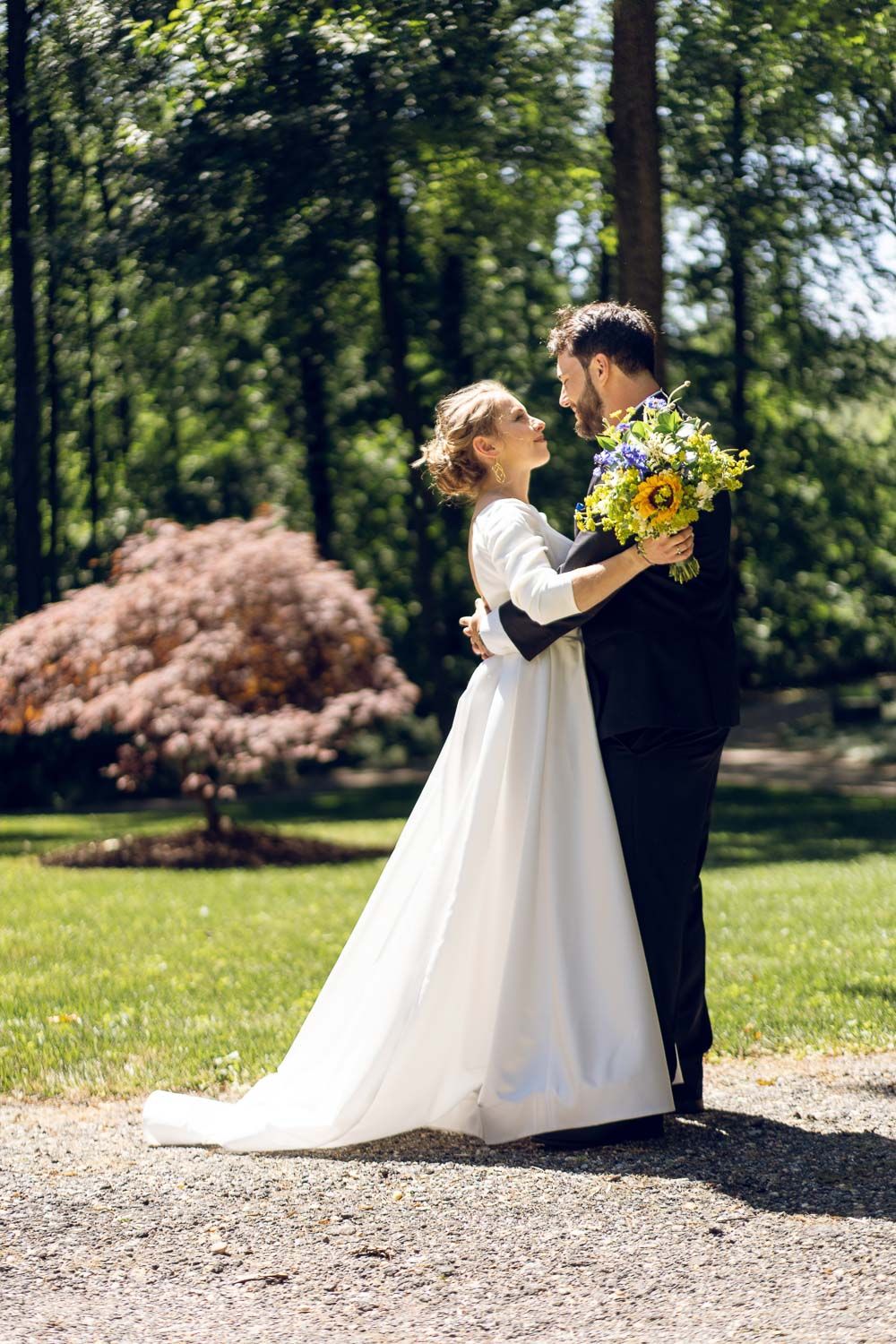 Bride and groom embracing face to face holding yellow and blue bouquet in sunlit park.
