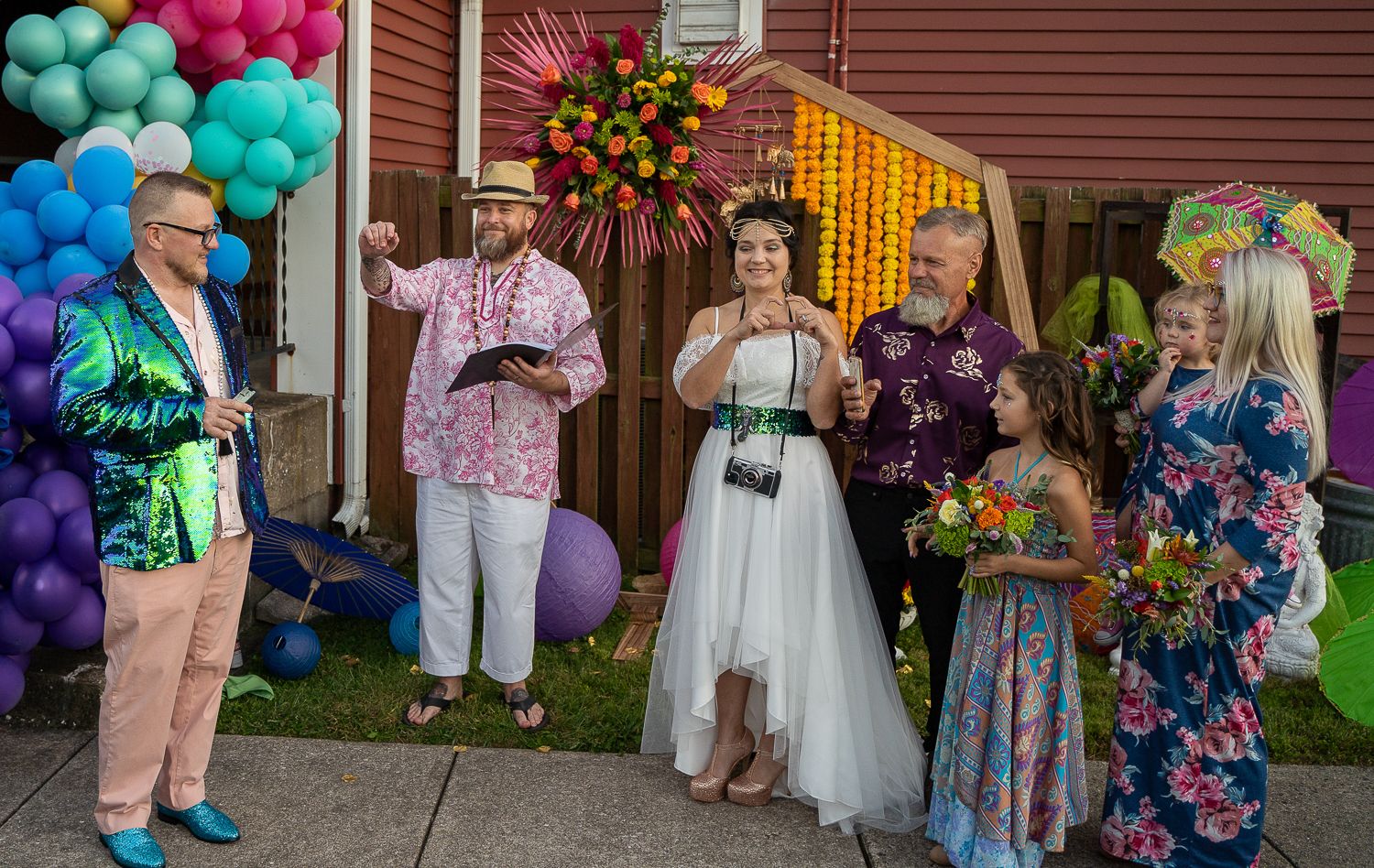 Colorful outdoor wedding ceremony with bride in white, officiant in hat, guests in vibrant attire and balloon decorations.