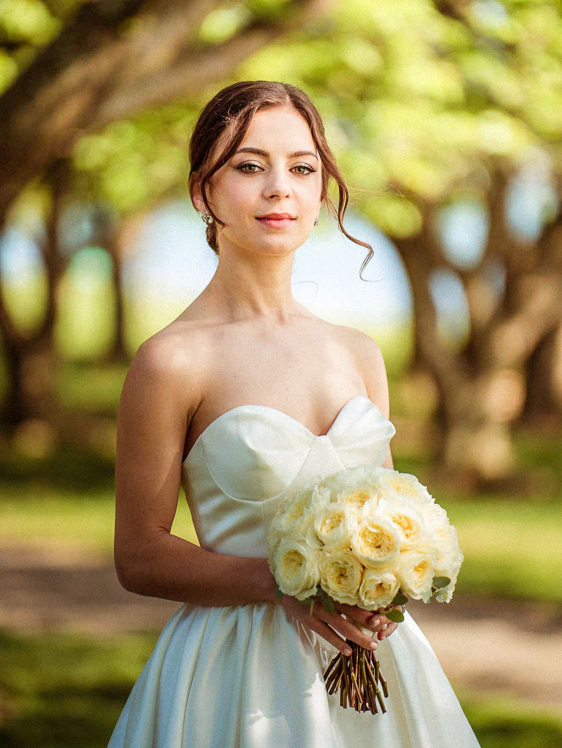 Beautiful bride in strapless gown holds white rose bouquet under sunlit trees.