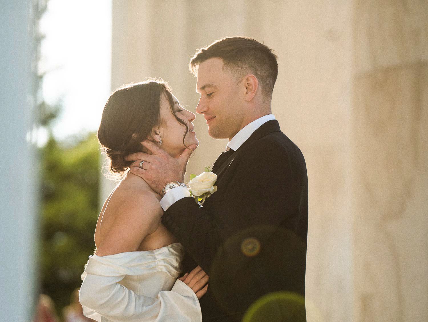 Bride and groom share a romantic almost-kiss bathed in golden sunlight near white columns.