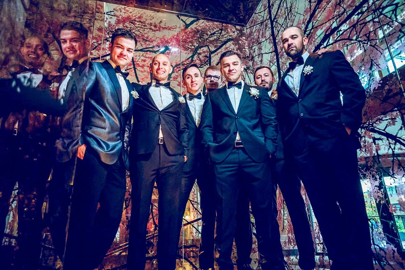 Groomsmen in black tuxedos smiling together in colorful artistic mirrored wedding reception venue.