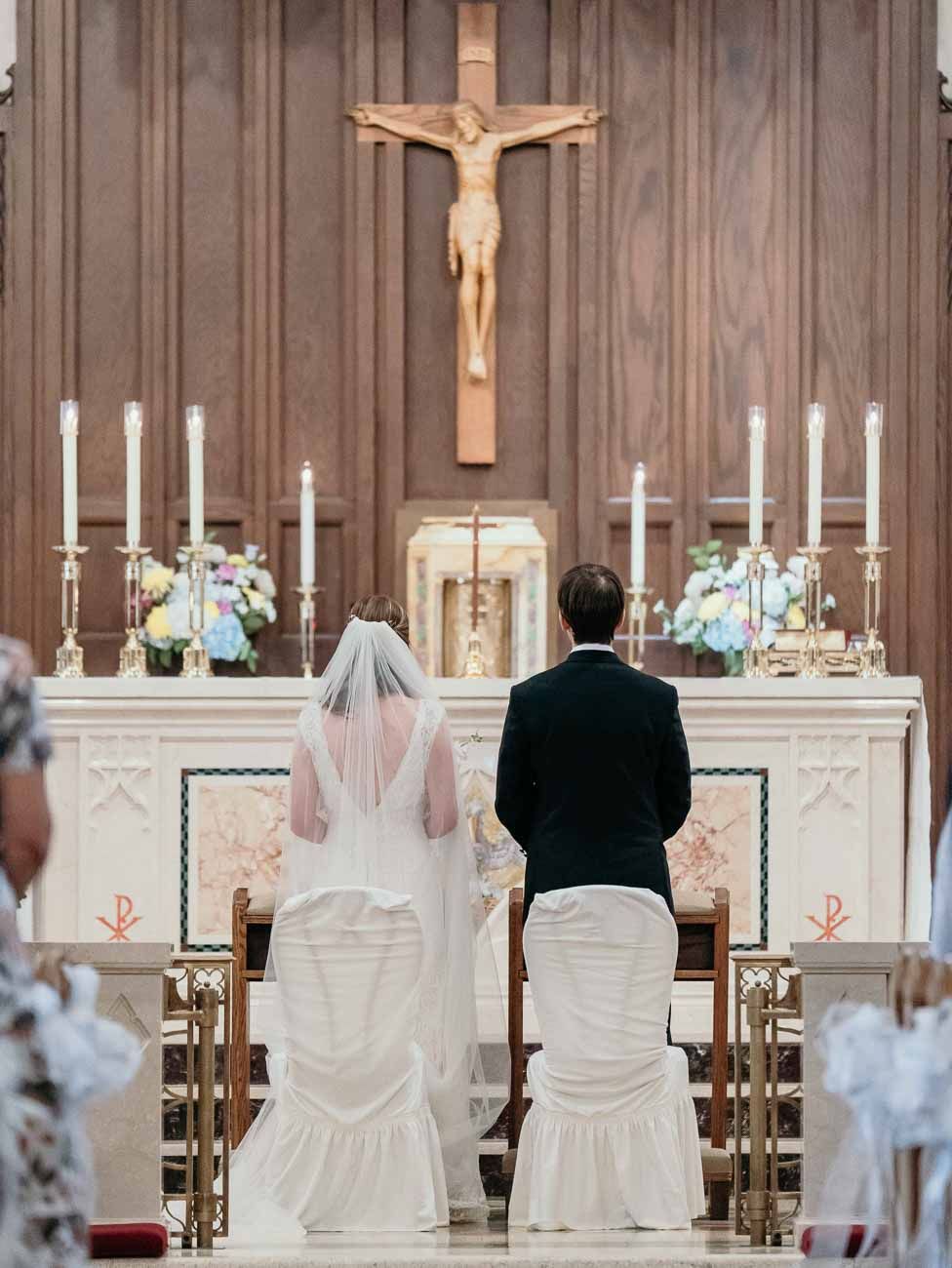 Bride and groom kneel together at ornate white altar beneath crucifix during wedding ceremony.