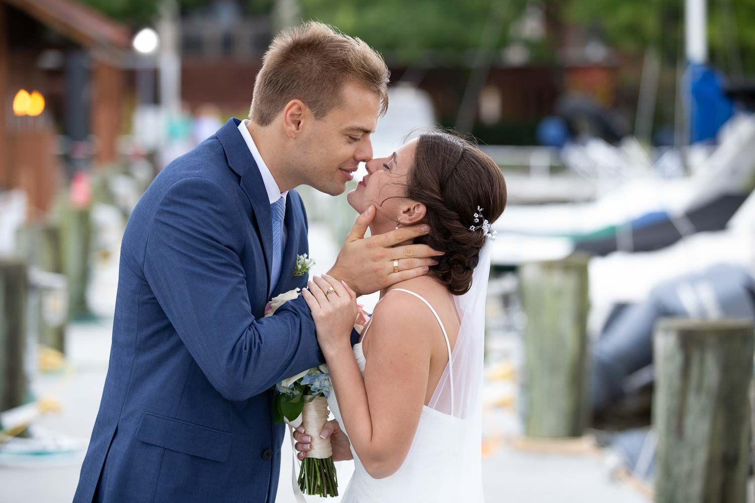 Bride and groom sharing tender moment at marina dock, groom in navy suit holding bouquet.