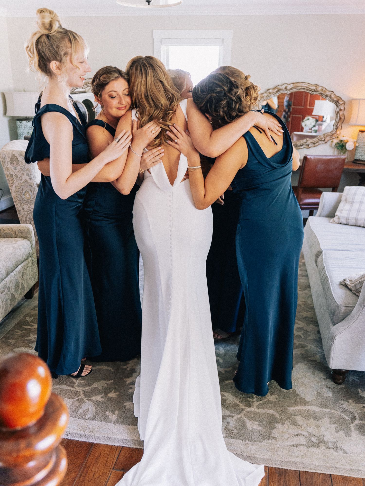 Bride in white gown embraced by bridesmaids in navy dresses during an emotional pre-wedding moment indoors.