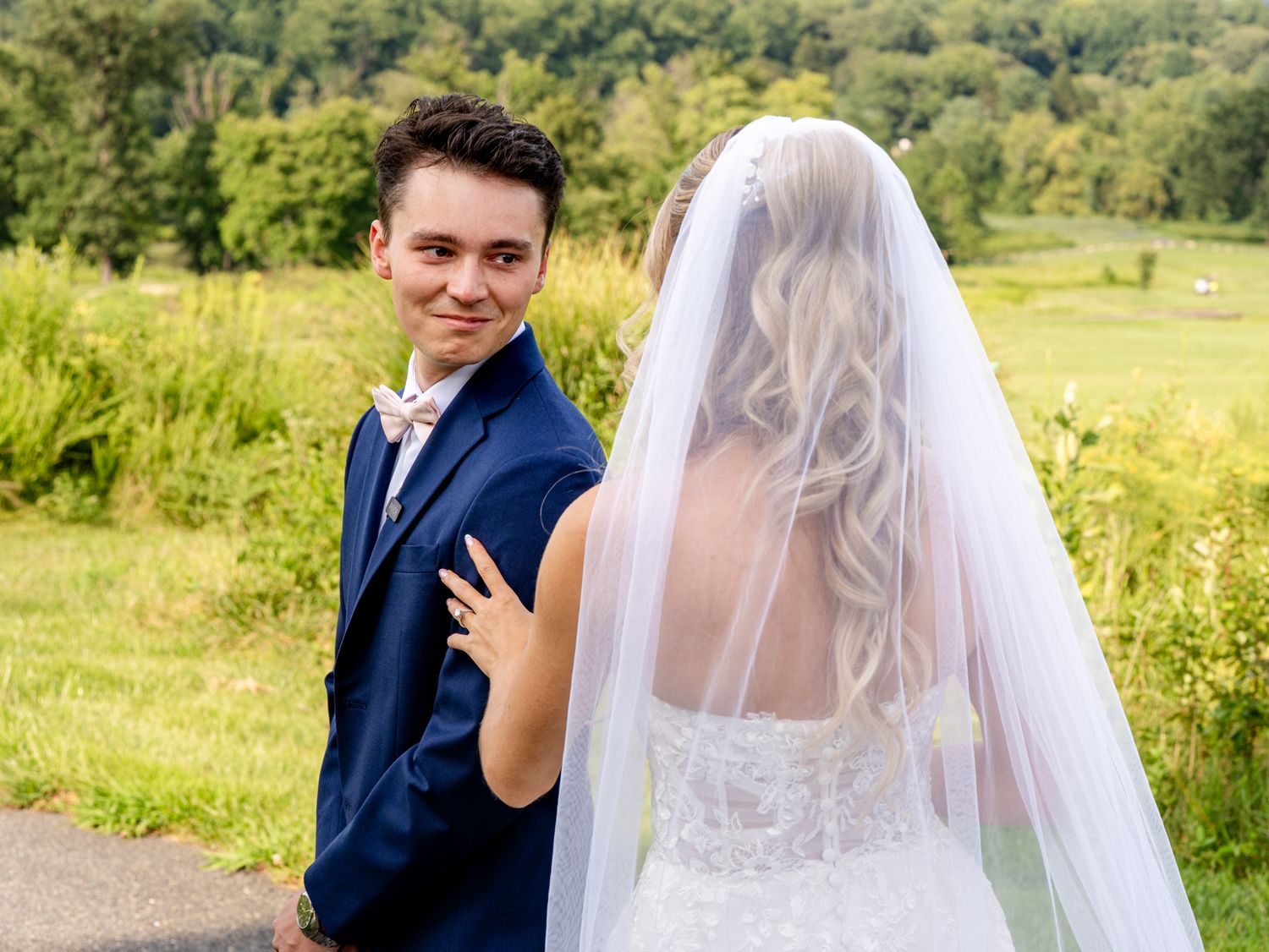 Groom in navy suit smiles at camera while bride in white lace gown and veil poses outdoors at wedding.