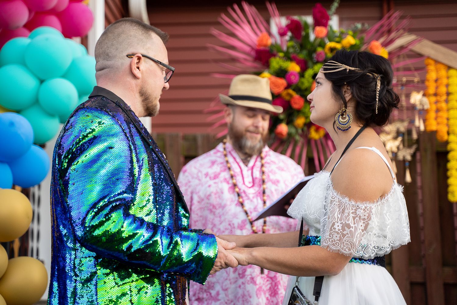 Couple exchanges rings at vibrant outdoor wedding with colorful balloons and tropical floral decorations.