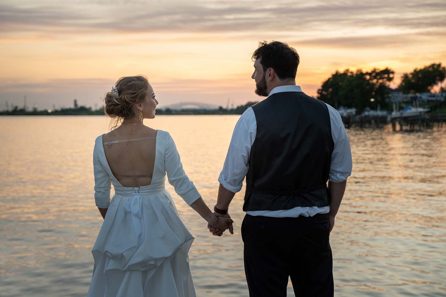Bride and groom holding hands watching sunset over calm water on their wedding day.