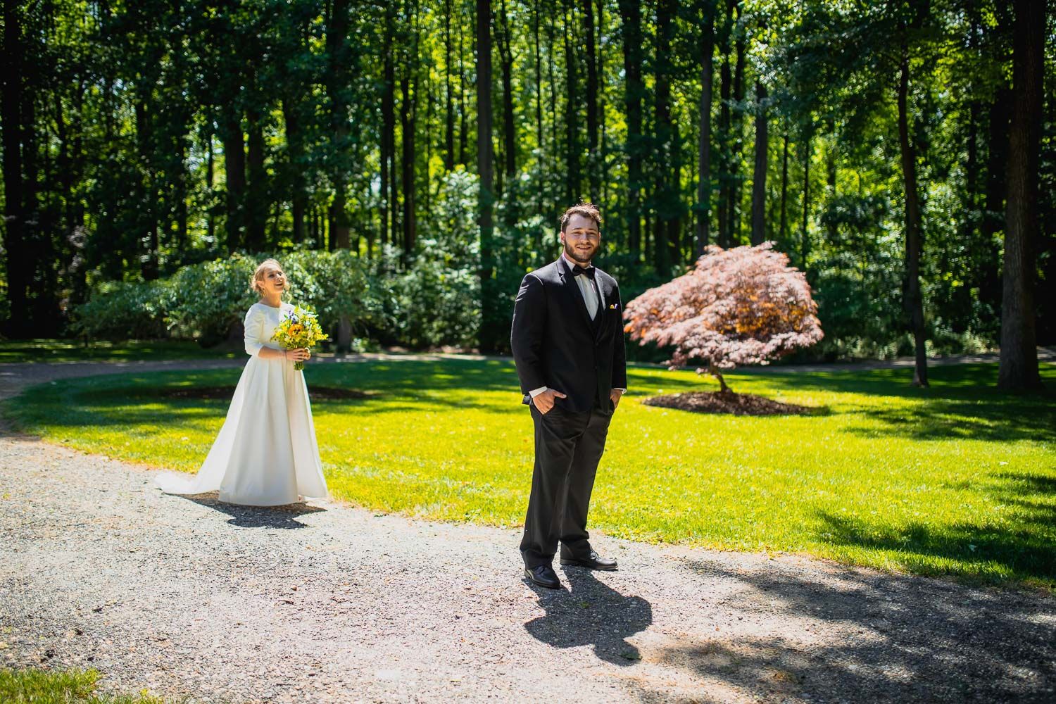 Bride and groom sharing first look moment on sunlit garden path surrounded by tall trees.