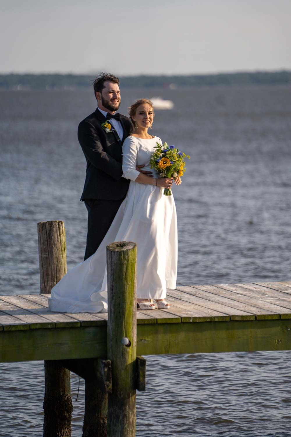 Smiling bride and groom posing together on a waterfront dock holding a sunflower bouquet.