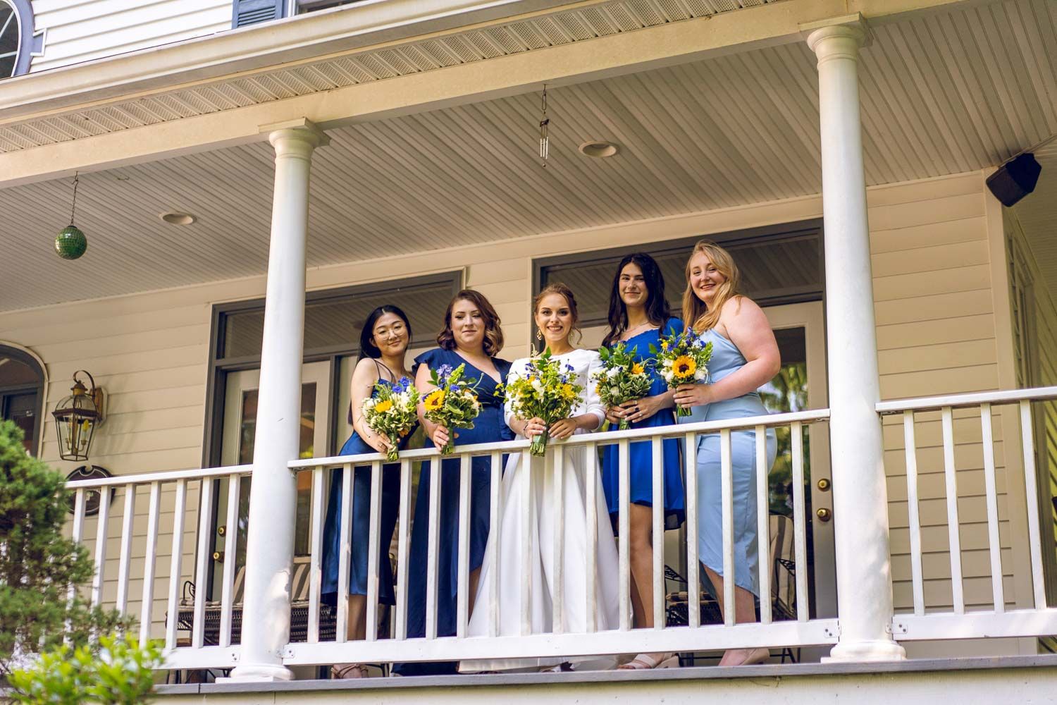 Four bridesmaids in blue dresses holding colorful bouquets pose on a white porch balcony.