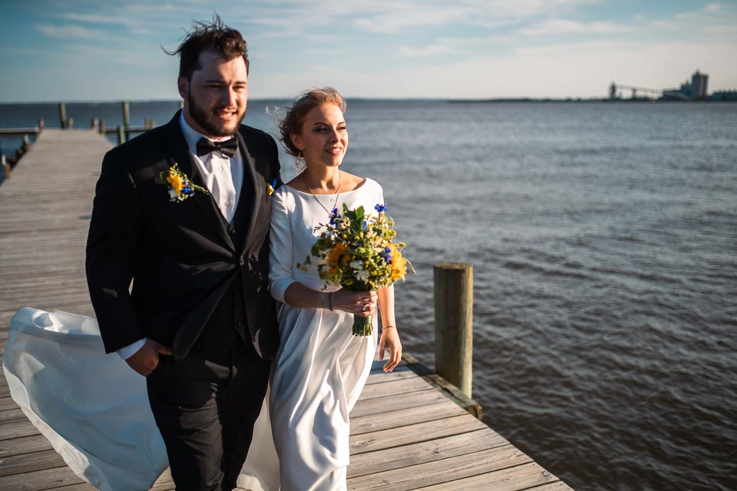 Bride and groom strolling on waterfront dock, brides veil blowing in wind, holding colorful wildflower bouquet.