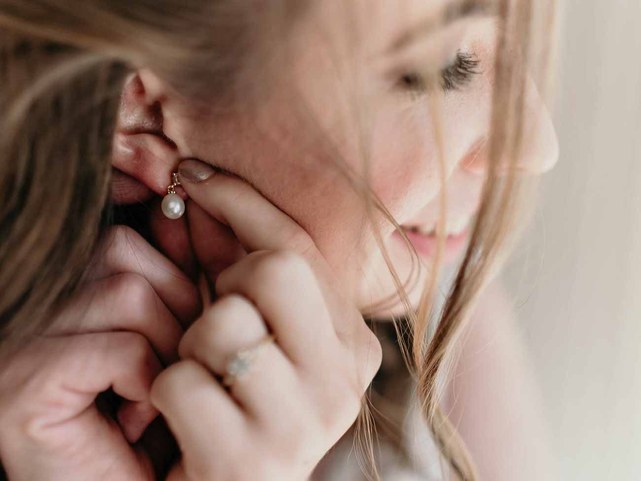 Close-up of bride putting on pearl drop earring with delicate ring on finger.