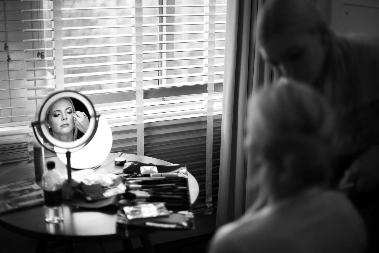Black and white photo of bride getting makeup done at vanity mirror.
