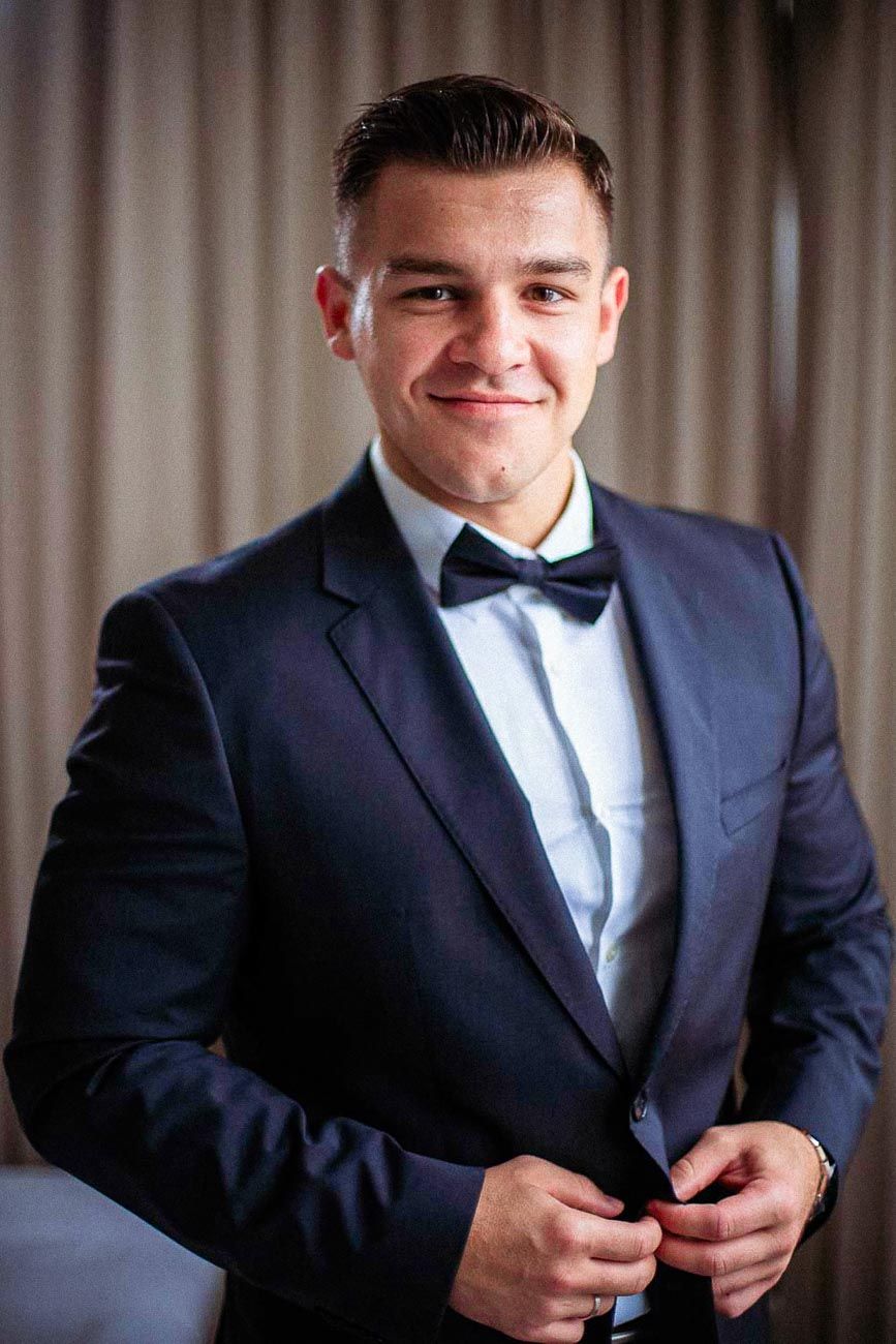 Smiling groom in navy tuxedo with black bow tie buttoning his jacket before wedding ceremony.