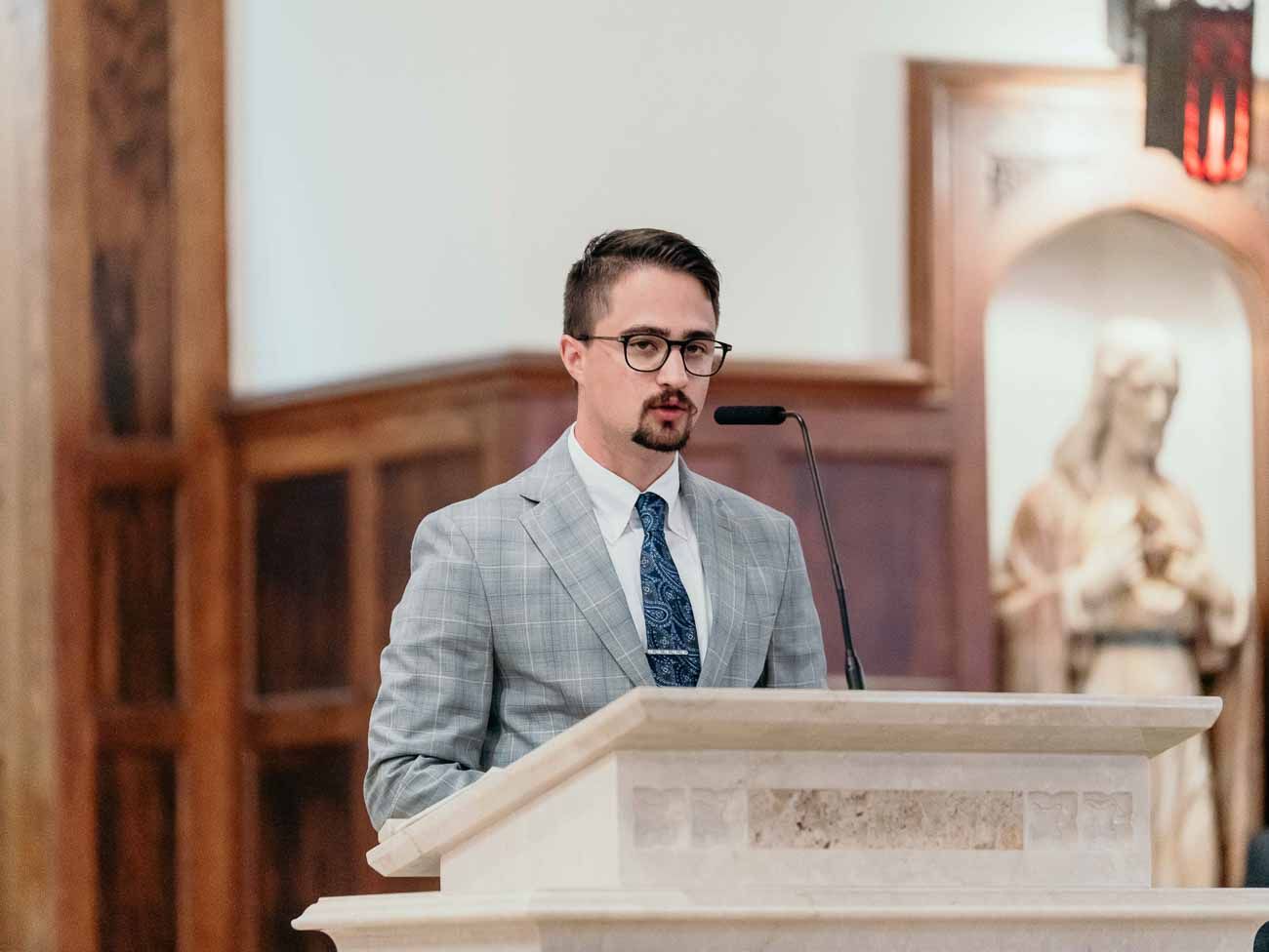 Man in gray suit speaking at a white marble church podium during a wedding ceremony.