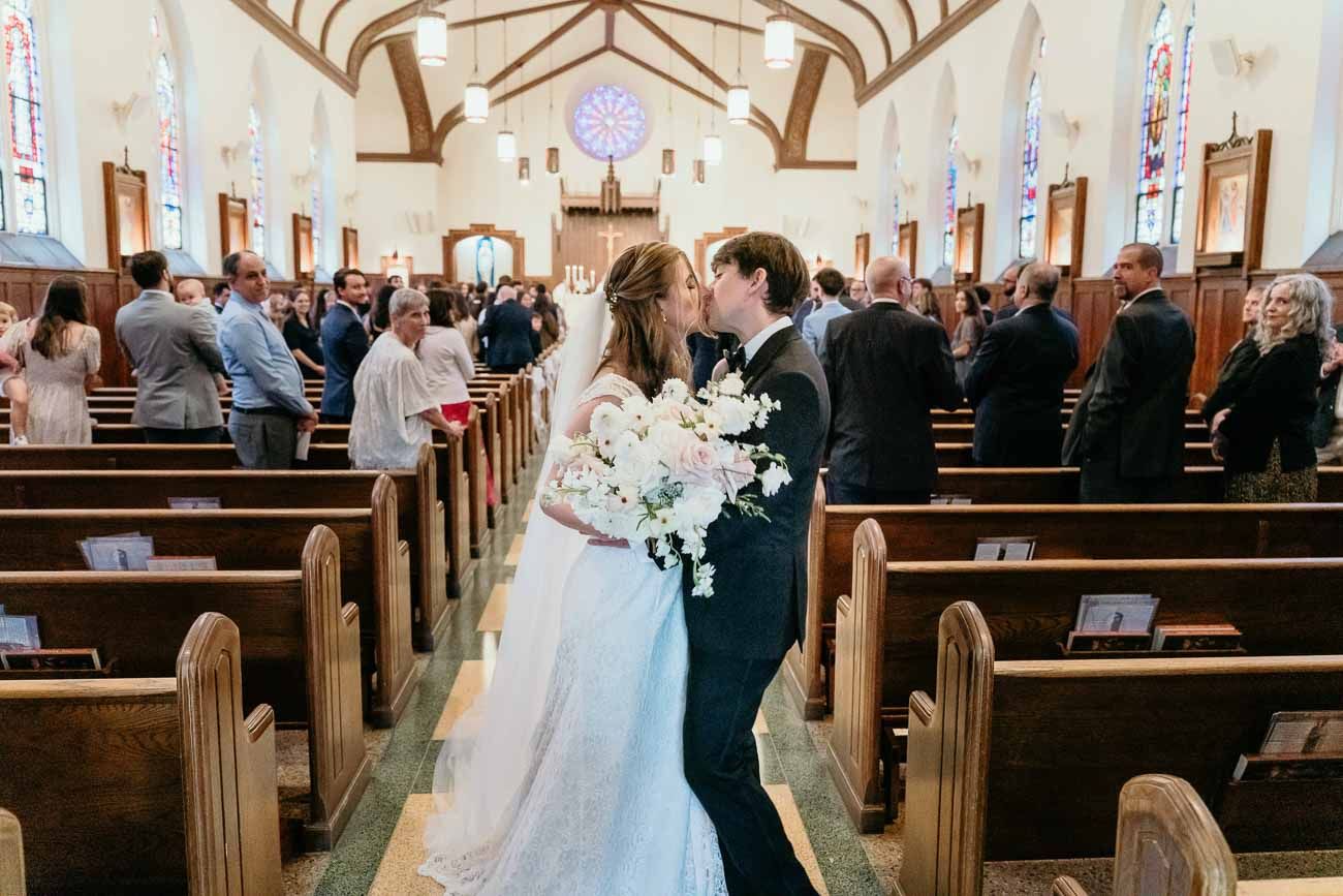 Newlyweds share first kiss in church aisle surrounded by guests after Catholic wedding ceremony.