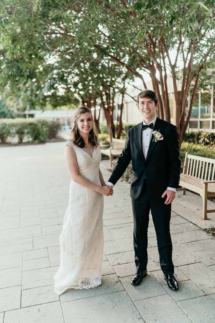 Smiling bride and groom holding hands and posing together under blooming trees on their wedding day.