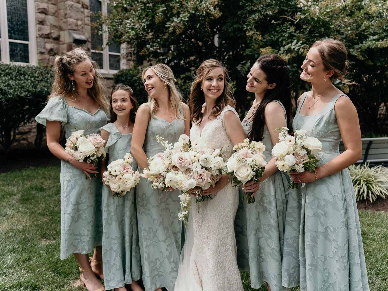 Bride in lace gown laughs with five bridesmaids in sage green dresses holding white bouquets.