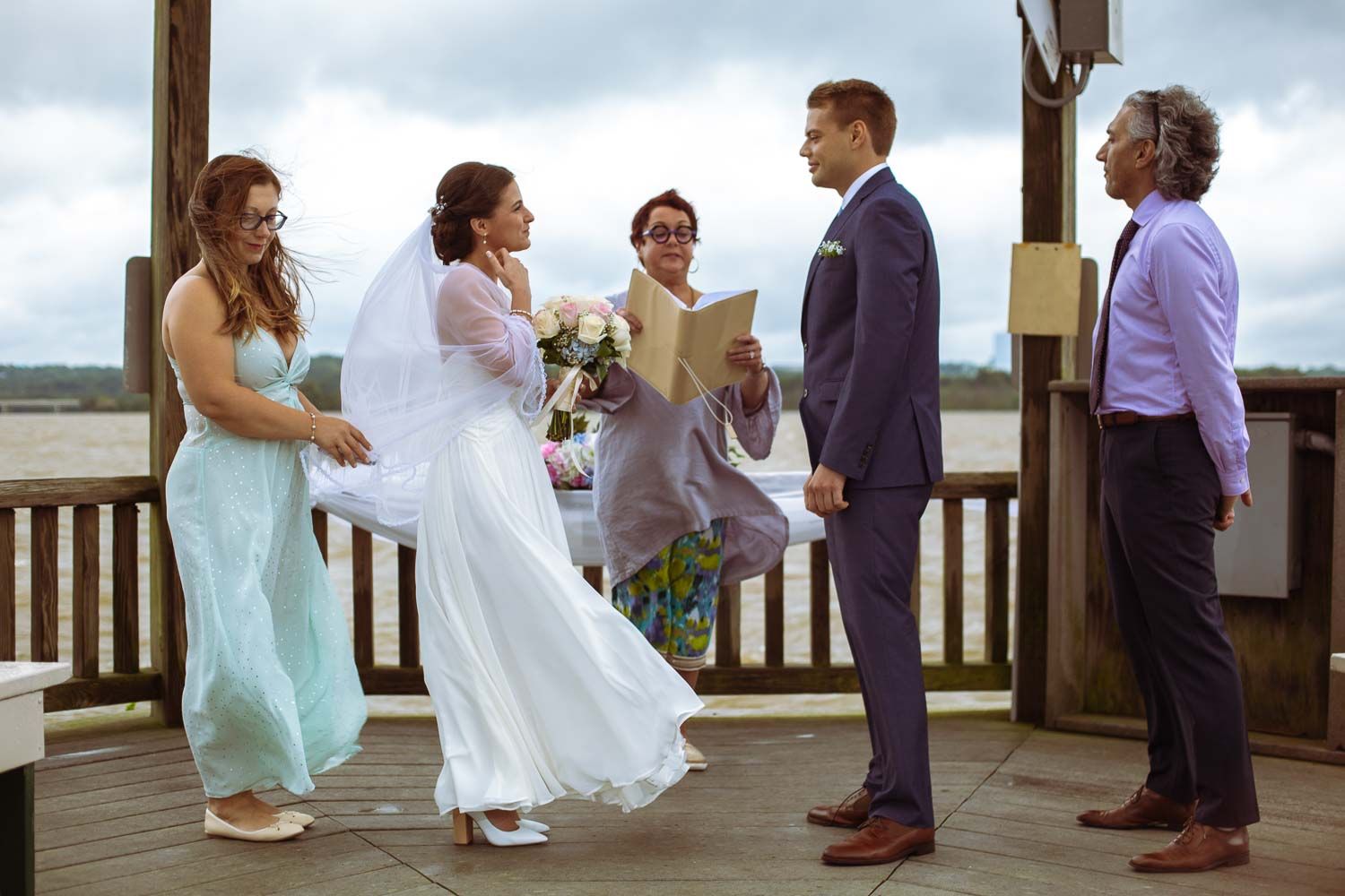Bride and groom exchange vows at outdoor waterfront wedding ceremony with officiant and wedding party on wooden deck.