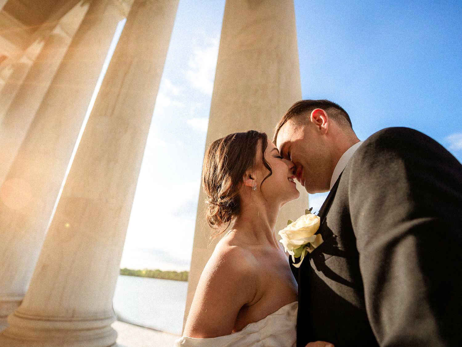 Bride and groom share a romantic moment between grand marble columns with sunlight glowing behind them.