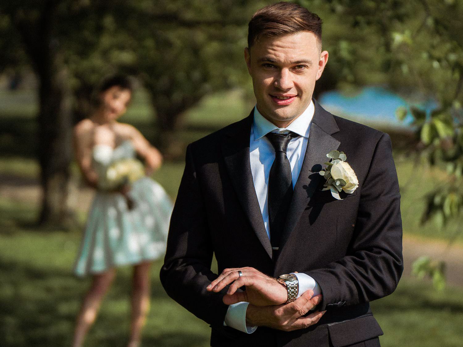 Groom in black suit adjusting cufflinks outdoors with bridesmaid in floral dress blurred in background.