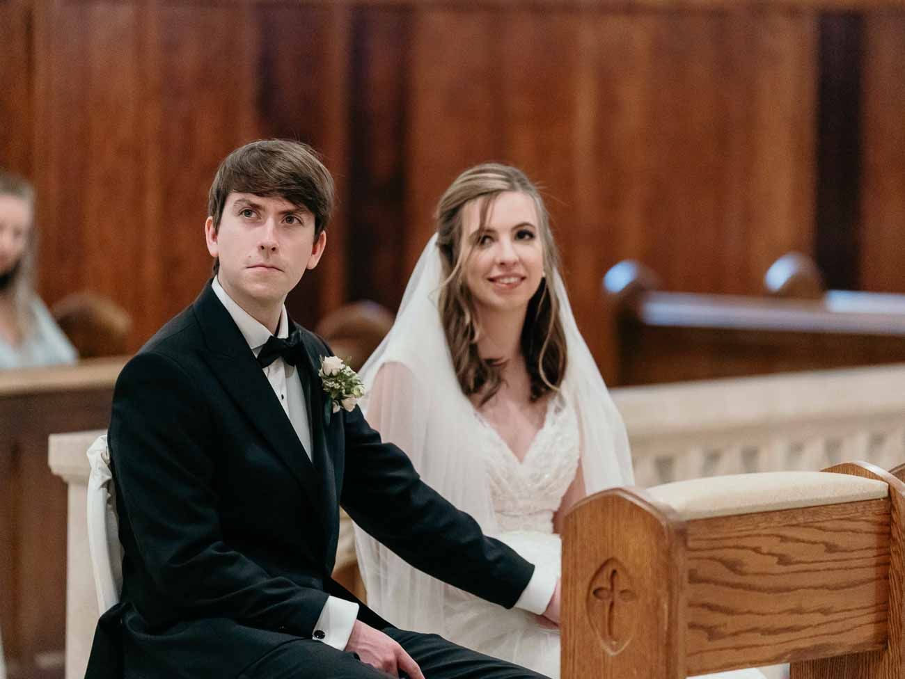 Bride in white veil and groom in black tuxedo seated together in a church pew during wedding.