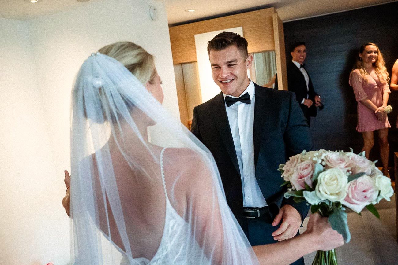 Groom in black tuxedo smiling at bride holding pink rose bouquet during first look reveal.