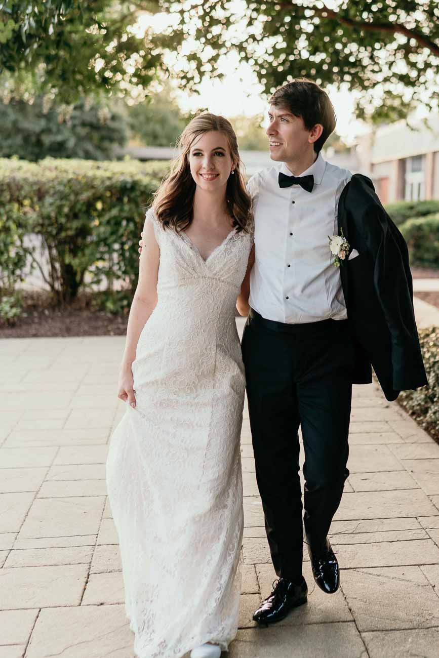 Newlyweds walk arm in arm outdoors, bride in lace gown and groom in black tuxedo with bow tie.