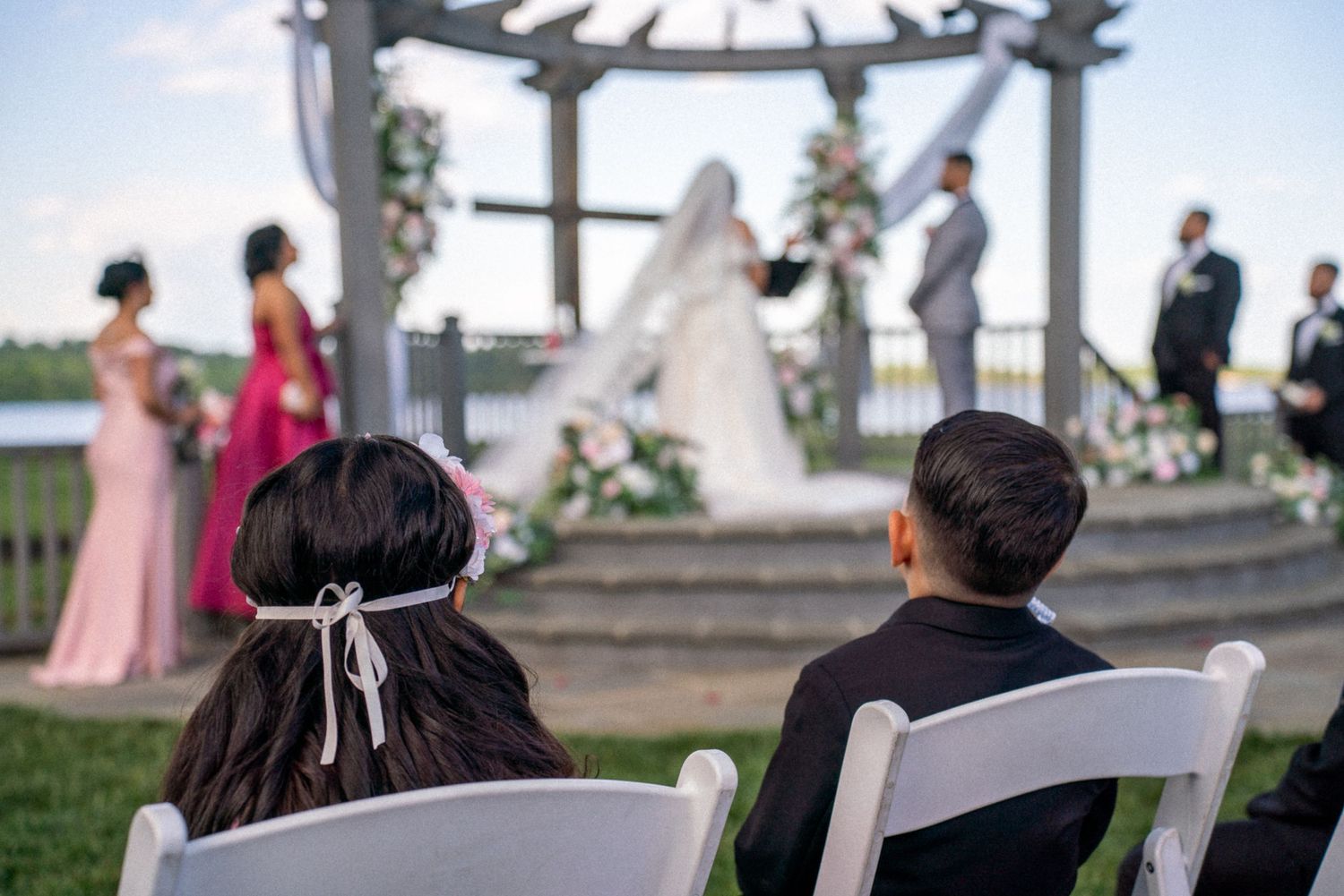 Guests seated at an outdoor wedding ceremony with bride and groom at a floral gazebo altar by a lake.